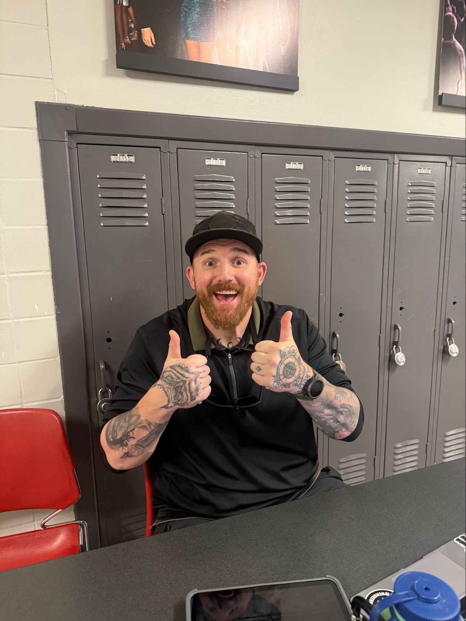 Graphic Design teacher Joe Zimka sits at a hall monitor post, displaying his tattoos. Photo courtesy of Brenna Chomicz, '29.